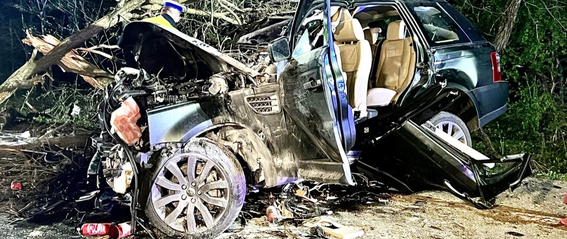 Na zdjęciu widać zniszczone auto osobowe koloru granatowego. Wnętrze auta jest koloru jasny beż. Za pojazdem widać drzewa i umundurowanego policjanta prowadzącego czynności dochodzeniowo – śledcze.