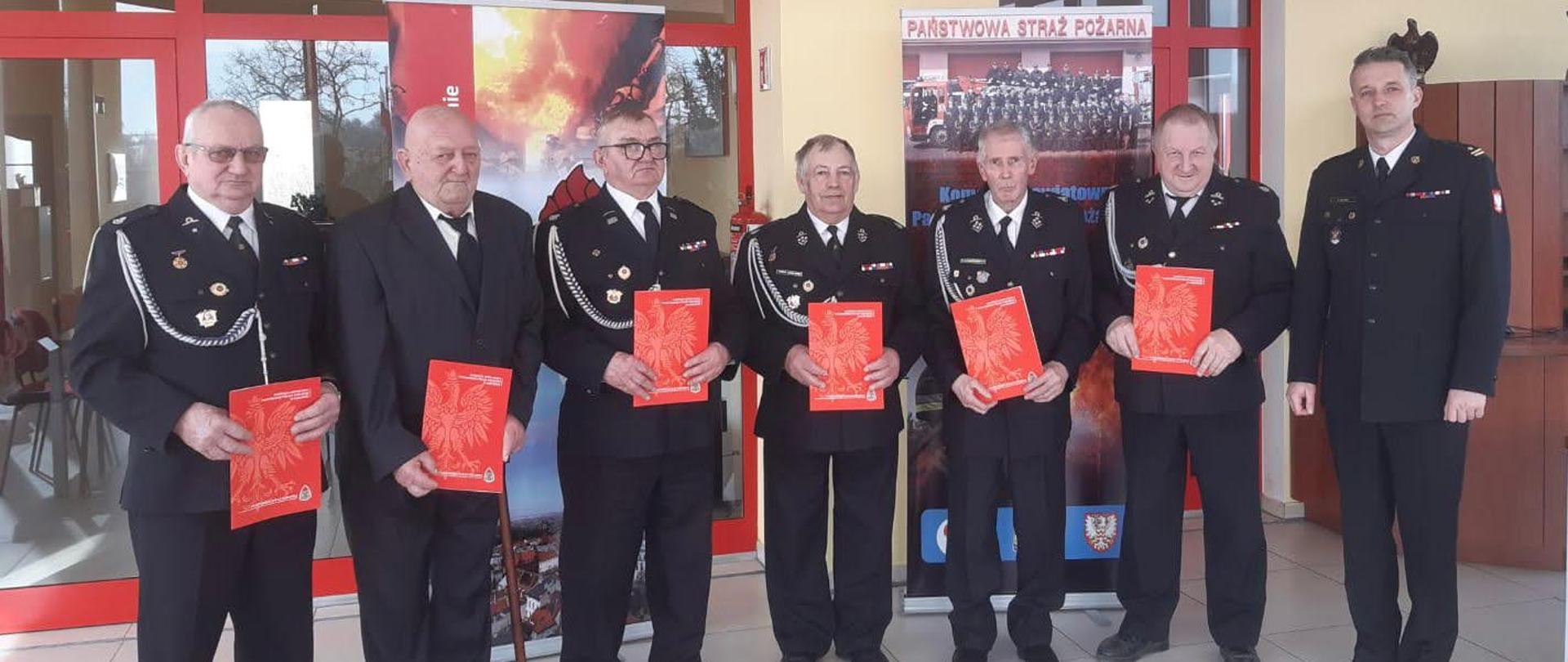 Na zdjęciu w sali sztabowej Komendy Powiatowej PSP w Chełmnie znajdują się druhowie, 6 osób wraz z Komendantem Powiatowym mł. bryg. Tomaszem Guzek, który wręcza druhom decyzje o przyznaniu świadczenia ratowniczego. Uczestnicy spotkania ustawili się do wspólnej fotografii.