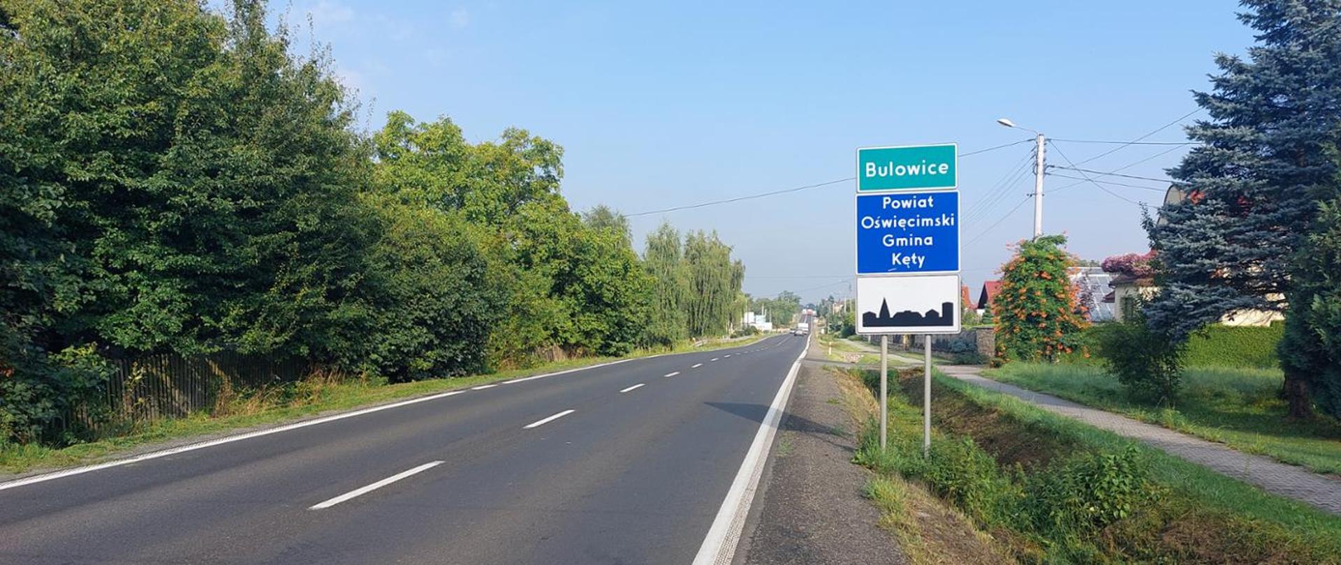 Na zdjęciu widoczna jest droga krajowa DK52 w miejscowości Bulowice. Centralnym elementem fotografii jest znak drogowy informujący o wjeździe do miejscowości Bulowice, która znajduje się w powiecie oświęcimskim, gminie Kęty. Droga jest asfaltowa, z wyraźnymi oznaczeniami pasów ruchu. Po lewej stronie drogi rozciąga się gęsty las, a po prawej widoczne są pojedyncze drzewa, krzewy oraz fragmenty zabudowań. Na niebie widnieje kilka chmur, a pogoda wydaje się być słoneczna.