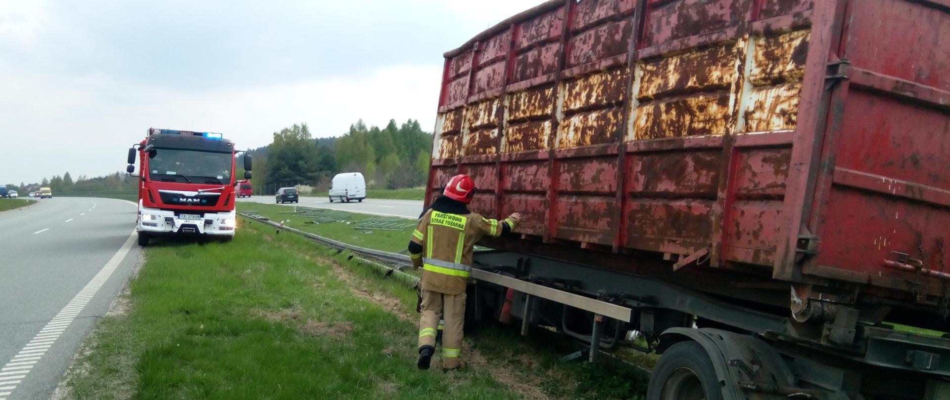 Na zdjęciu przyczepa ze złomem po wjechaniu w bariery ochronne i stojący przy niej strażak. Na pasie zieleni elementy zniszczonej bariery, a po lewej stronie stoi zaparkowany samochód pożarniczy