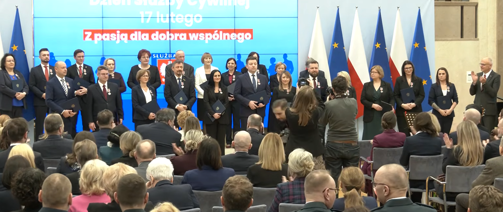 Na zdjęciu widać uroczystość oficjalną w sali konferencyjnej. Na scenie stoi grupa osób w strojach formalnych, część z nich trzyma teczki i ma przypięte odznaczenia. W tle znajdują się flagi Polski i Unii Europejskiej oraz ekran z napisem o Dniu Służby Cywilnej. Przed sceną siedzi publiczność, a kilka osób robi zdjęcia