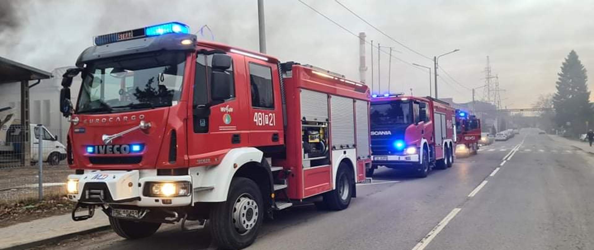 Zdjęcie obrazuje stojące na ulicy Mościckiego w Skarżysku-Kamiennej trzy zastępy z Państwowej Straży Pożarnej. Z lewej strony widać unoszący się dym z hali magazynowej.