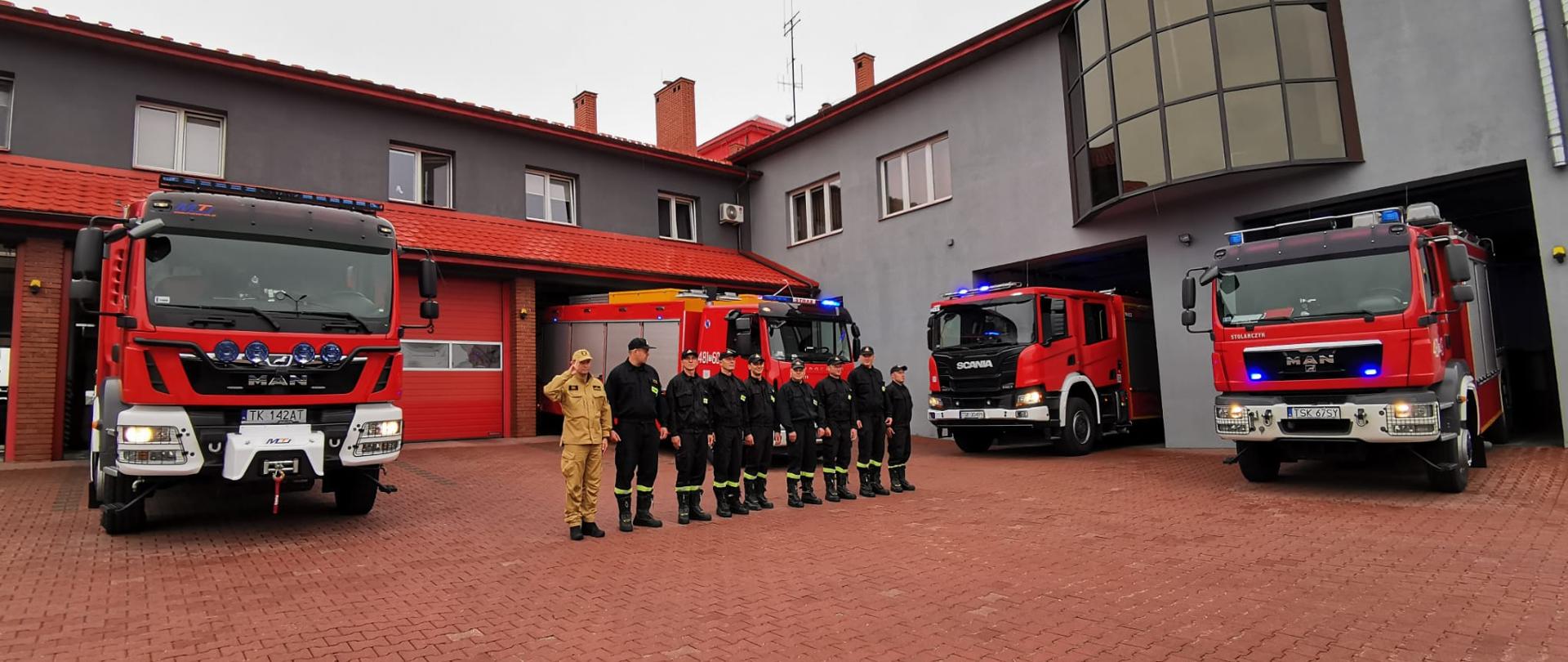 Na zdjęciu widać strażaków KP PSP Skarżysko-Kamienna ubranych w ubrania koszarowe, oddających hołd Powstańcom Warszawskim. Stoją oni przed garażami, są wyprowadzone również samochody bojowe z włączonymi sygnałami świetlnymi i dźwiękowymi. W tle widać budynek Komendy Powiatowej. 