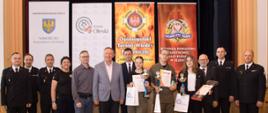 W sali w Miejskim Domu Kultury w Oleśnie są ustawieni w szeregu laureaci III kategorii wiekowej etapu powiatowego OTWP, organizatorzy i zaproszeni goście. W tle stoją 4 banery: Samorządu Województwa Opolskiego, Starostwa Powiatowego, OTWP, Komendy Powiatowej Państwowej Straży Pożarnej w Oleśnie.