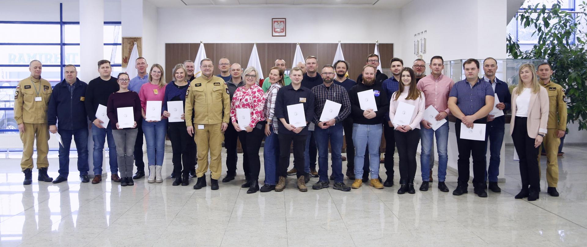 Pamiątkowe zdjęcie uczestników szkolenia (25 osób) w towarzystwie Zastępcy Komendanta Centralnj Szkoły PSP, dwóch funkcjonariuszy CS PSP oraz kobiety