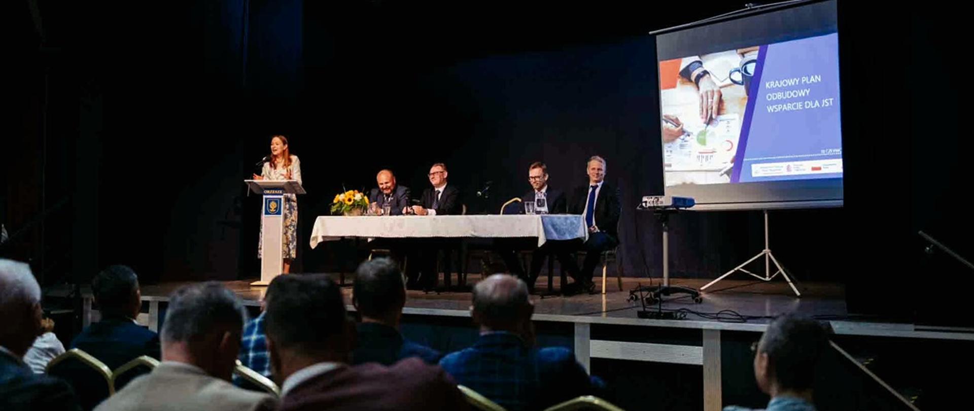 Minister funduszy i polityki regionalnej Katarzyna Pełczyńska-Nałęcz stoi na podium przy mównicy i przemawia, po jej lewej stronie stoi stół przy którym siedzą dyrektorzy departamentów w MFiPR, na pierwszym planie widzimy uczestników spotkania siedzących w fotelach
