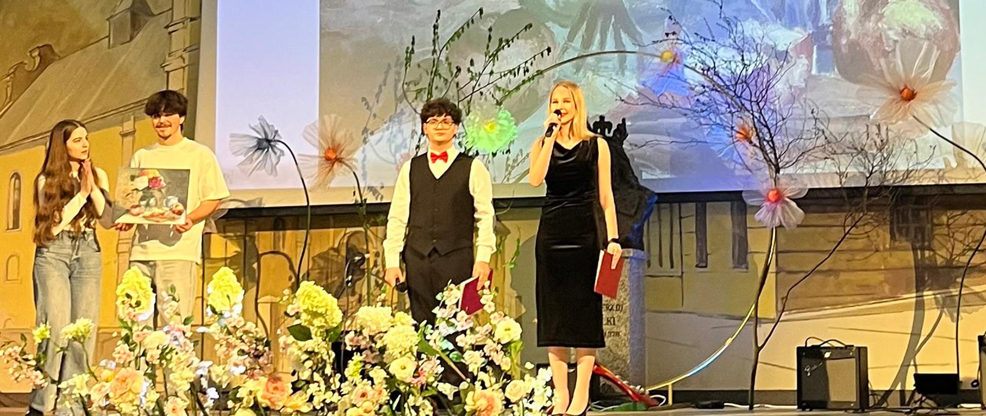 Zdjęcie przedstawia moment na scenie, gdzie czwórka młodych osób stoi obok siebie w uroczystych pozach. Po lewej stronie para nastolatków wspólnie trzyma niewielki obraz z motywem kwiatowym; dziewczyna bije brawo, a chłopak uśmiecha się do widowni. Pośrodku stoi chłopak w eleganckiej kamizelce, a po prawej kobieta w czarnej sukni trzyma mikrofon i czerwoną teczkę. Całość odbywa się w uroczystej atmosferze, podkreślonej przez kwiatowe dekoracje i monumentalną projekcję obrazu na ścianie za nimi.