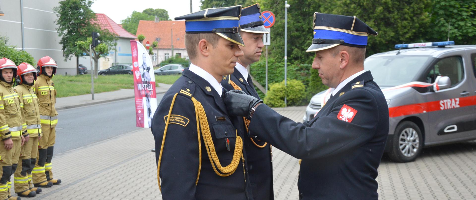 strazak w ciemnym mundurze w rogatywce przypina medal drugiemu strażakowi do munduru, w tle samochód strażacki i strażacy w mundurach bojowych i hełmach