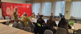 Spotkanie strażaków Państwowej Straży Pożarnej w sali konferencyjnej. Jeden z uczestników stoi i przemawia, trzymając kartkę, a pozostali siedzą przy stołach i słuchają. W tle widoczna jest czerwona ścianka z godłem Polski i napisem PSP, a na stołach stoją butelki z wodą.