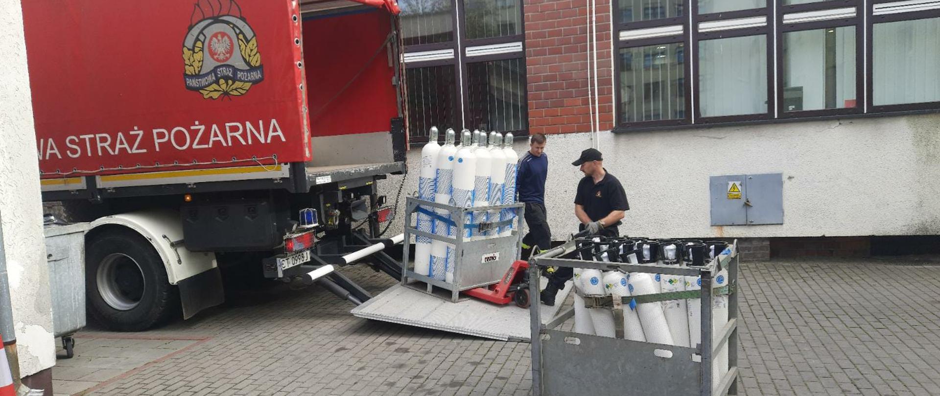 Na zdjęciu widać dwóch strażaków, którzy rozładowują skrzynie z butlami z tlenem. Strażacy znajdują się przed szpitalem.