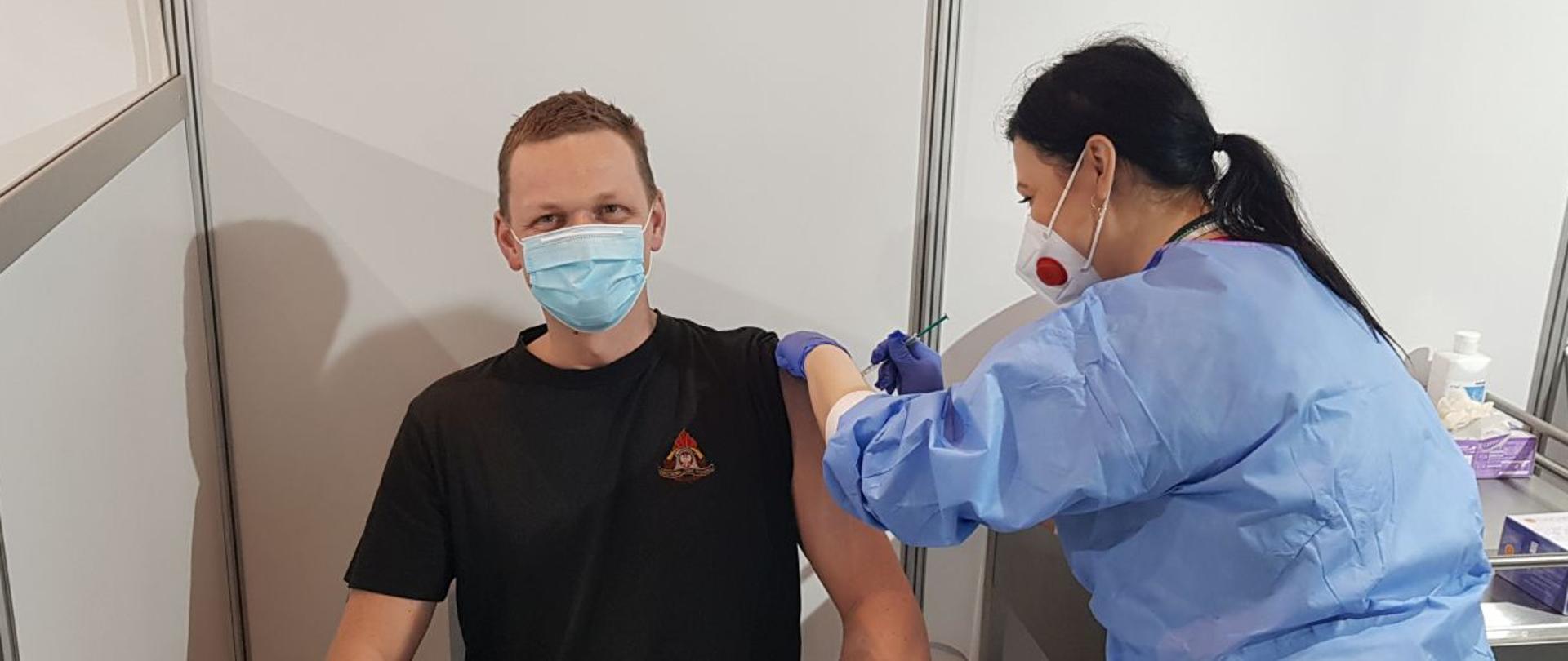 Zdjęcie przedstawia funkcjonariusza KM PSP w Jaworznie Michała Dyla w czasie wykonywania szczepienia przeciwko Covid 19 przez pielęgniarkę.