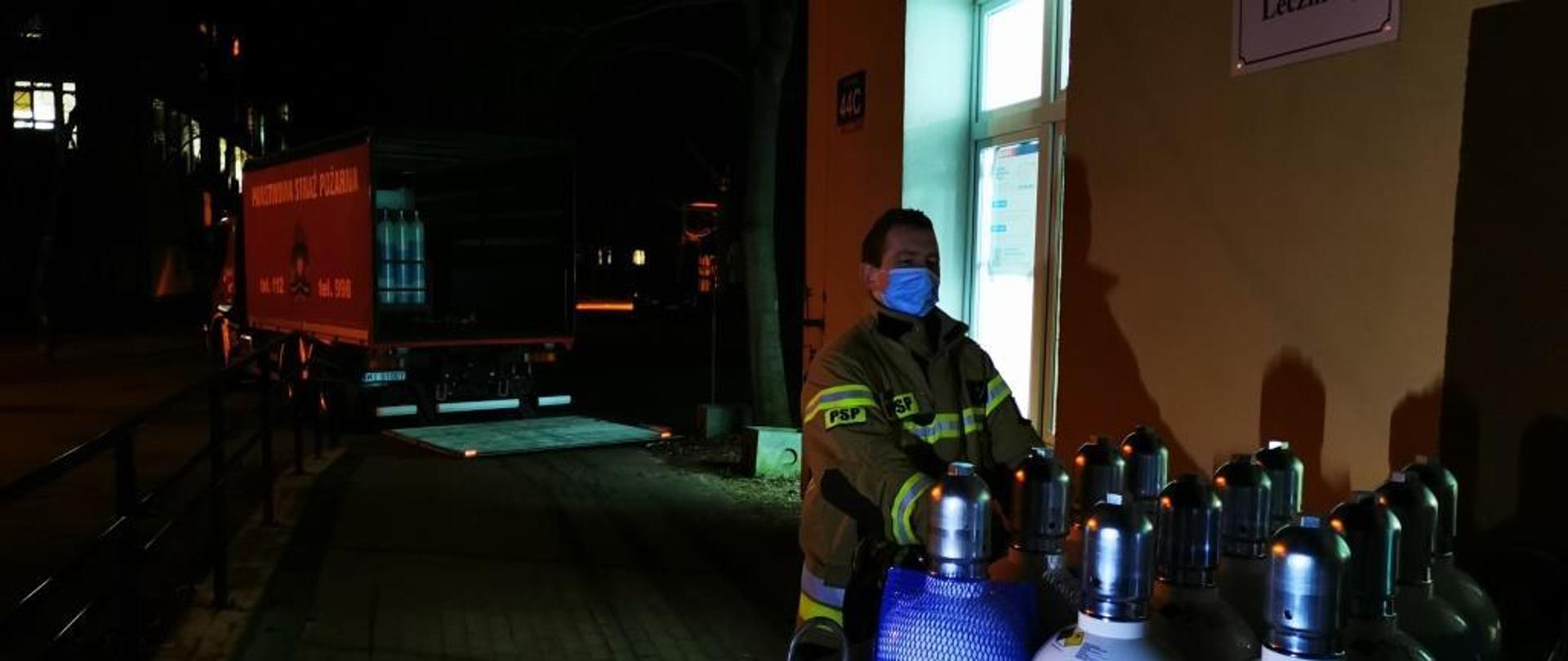 Zdjęcie przedstawia strażaka przewożącego na wózku butle wypełnione tlenem medycznym do szpitala. Strażak ubrany w ubranie specjalne w kolorze żółtym z elementami odblaskowymi. Na twarzy posiada również maseczkę ochronną. Po lewej stronie w oddali widać pojazd ciężarowy na którym załadowane znajdują się jeszcze butle z tlenem. Zdjęcie wykonane w porze nocnej. 