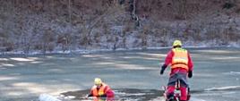 Na zdjęciu dwóch ratowników przeprowadza ćwiczenie ratownictwa wodno-lodowego na zamarzniętym jeziorze. Jeden z ratowników w kamizelce ratunkowej jest w przeręblu. Po prawej stronie jest ratownik w kamizelce i kasku, przywiązany do linii bezpieczeństwa, podąża w kierunku przerębla, gotowy do udzielenia pomocy. Cała dwójka jest odpowiednio wyposażona w specjalistyczny sprzęt do ćwiczeń na lodzie. Zdjęcie pokazuje praktyczne metody działania, które są stosowane podczas rzeczywistych akcji ratunkowych na lodzie.
