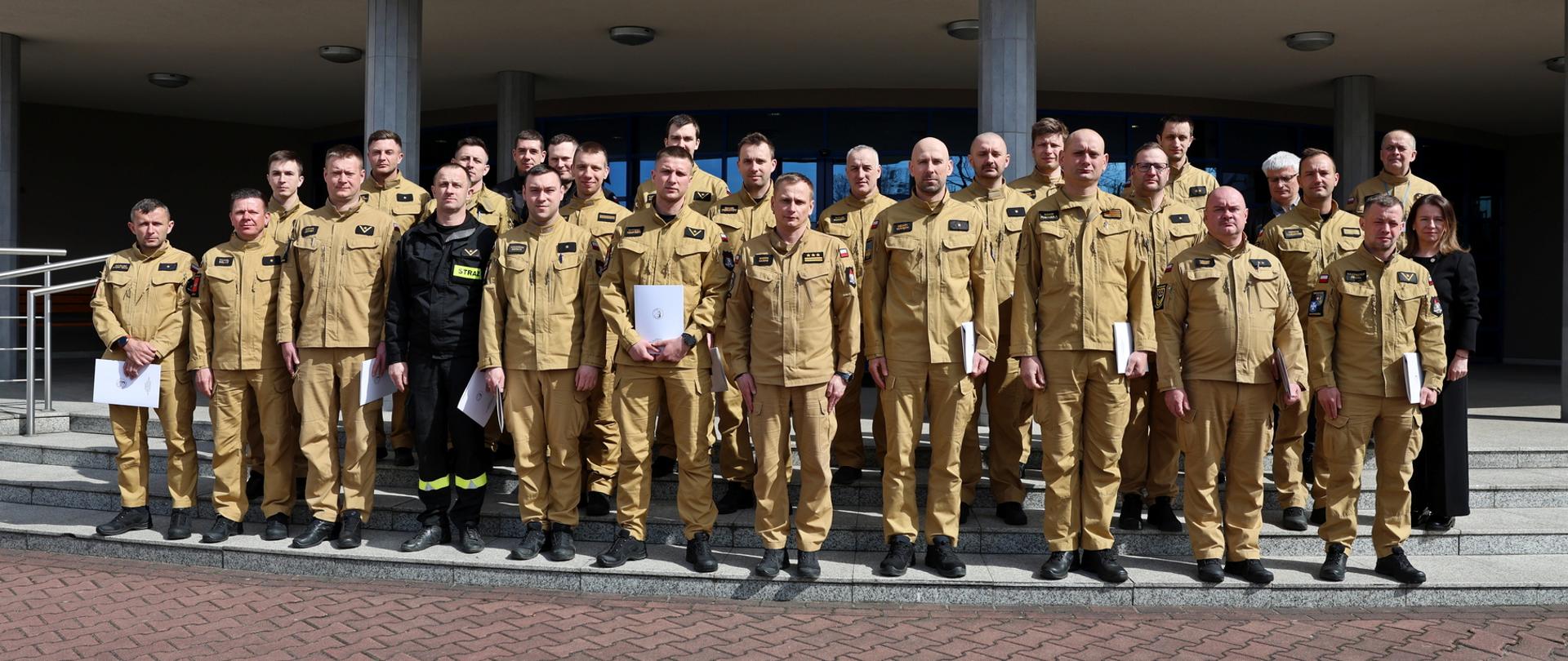 Pamiątkowe zdjęcie grupowe funkcjonariuszy PSP uczestniczących w szkoleniu z zakresu awaryjnego rozładunku cystern (24 osoby) wykonane przed budynkiem. Wśród uczestników na zdjęciu są również: Zastępca Komendanta Szkoły, kobieta, funkcjonariusz CS PSP oraz mężczyzna będący instruktorem podczas zajęć