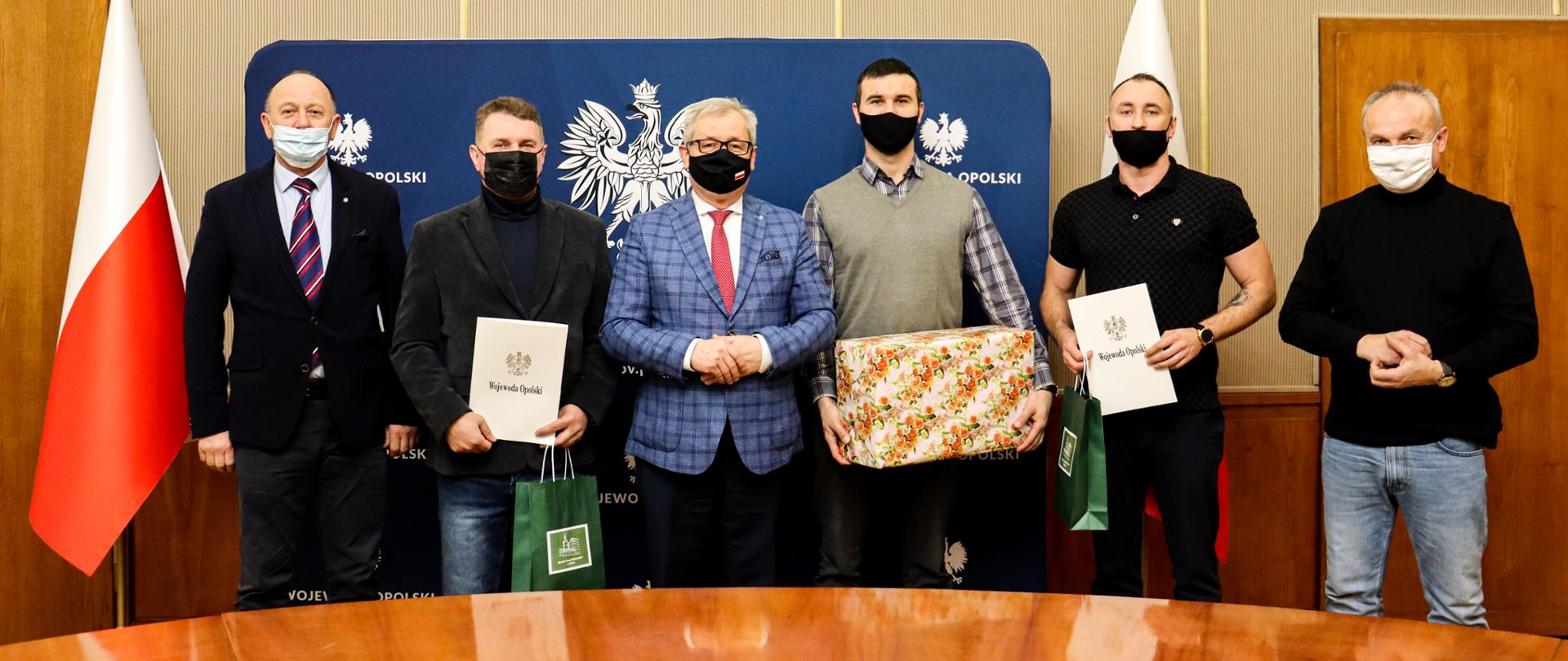Na zdjęciu 6 mężczyzn stojących na tle ścianki z napisem i logo Wojewoda Opolski. Wojewoda w środku. Panowie pozują do zdjęcia. Jeden z mężczyzn trzyma w dłoni paczkę z prezentem, dwóch pozostałych w dłoniach trzymają białą teczkę z orłem. Po lewej stronie kadru widać biało-czerwoną flagę. 