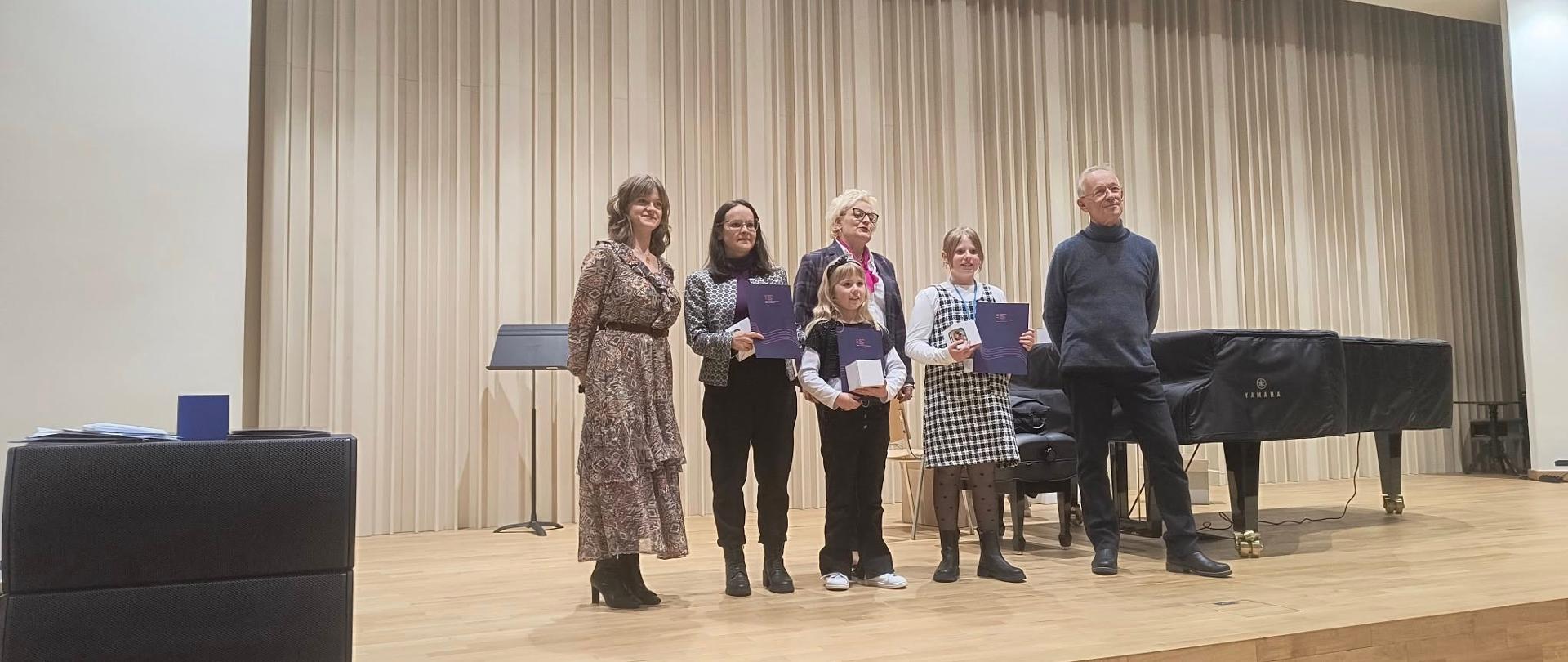 Na jasnej scenie stoją uczestnicy warsztatów wraz z opiekunami. W dłoniach trzymają dyplomy.