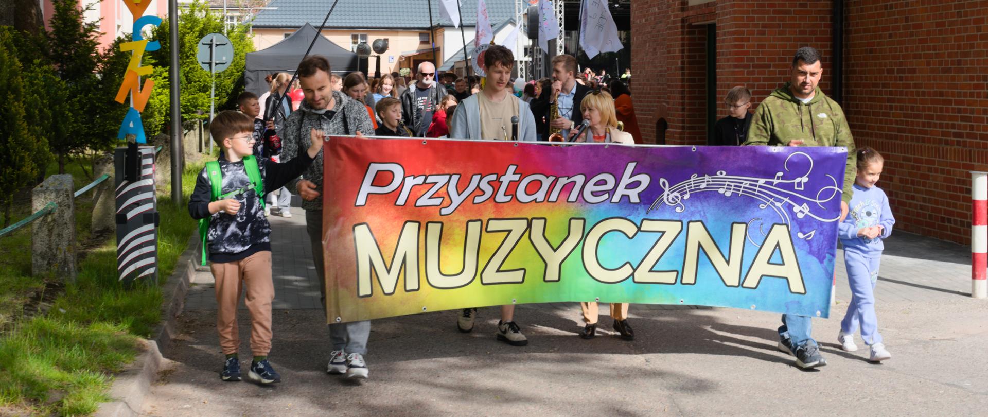 Na zdjęciu widać kolorowy pochód ludzi niosących transparent z napisem „Przystanek MUZYCZNA” w tęczowych barwach i nutami. Uczestnicy marszu, w tym dzieci i dorośli, maszerują uliczką między budynkami, trzymając flagi. Po lewej stronie stoi pionowy napis „PRZYSTANEK MUZYCZNA” z kolorowych liter. W tle widać scenę, co sugeruje, że wydarzenie ma charakter festiwalu muzycznego lub parady.