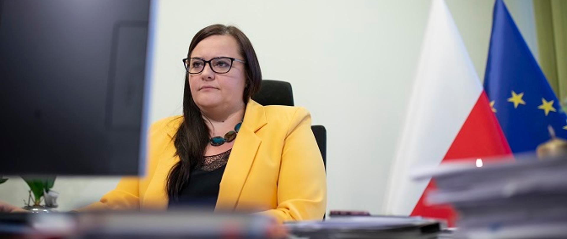 Minister Małgorzata Jarosińska-Jedynak siedzi przy komputerze za biurkiem, W tle flagi Polski i UE.