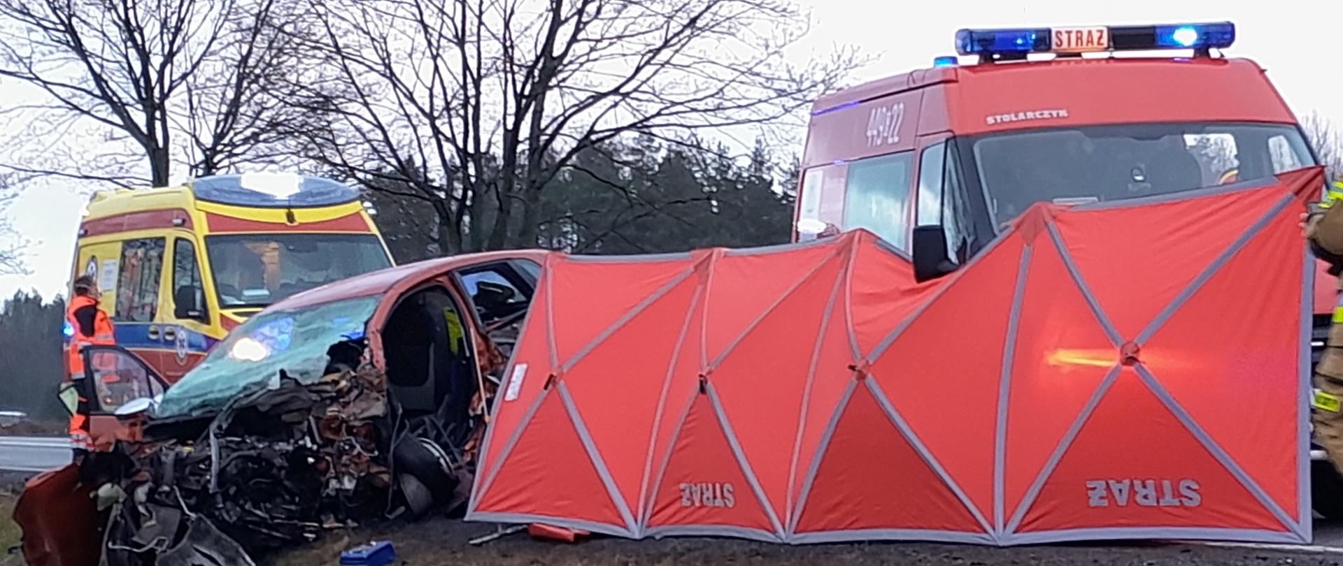 Miejsce wypadku osłonięte czerwonym parawanem. Z lewej strony znajduje się uszkodzony samochód osobowy. W tle karetka pogotowia oraz samochód strażacki.