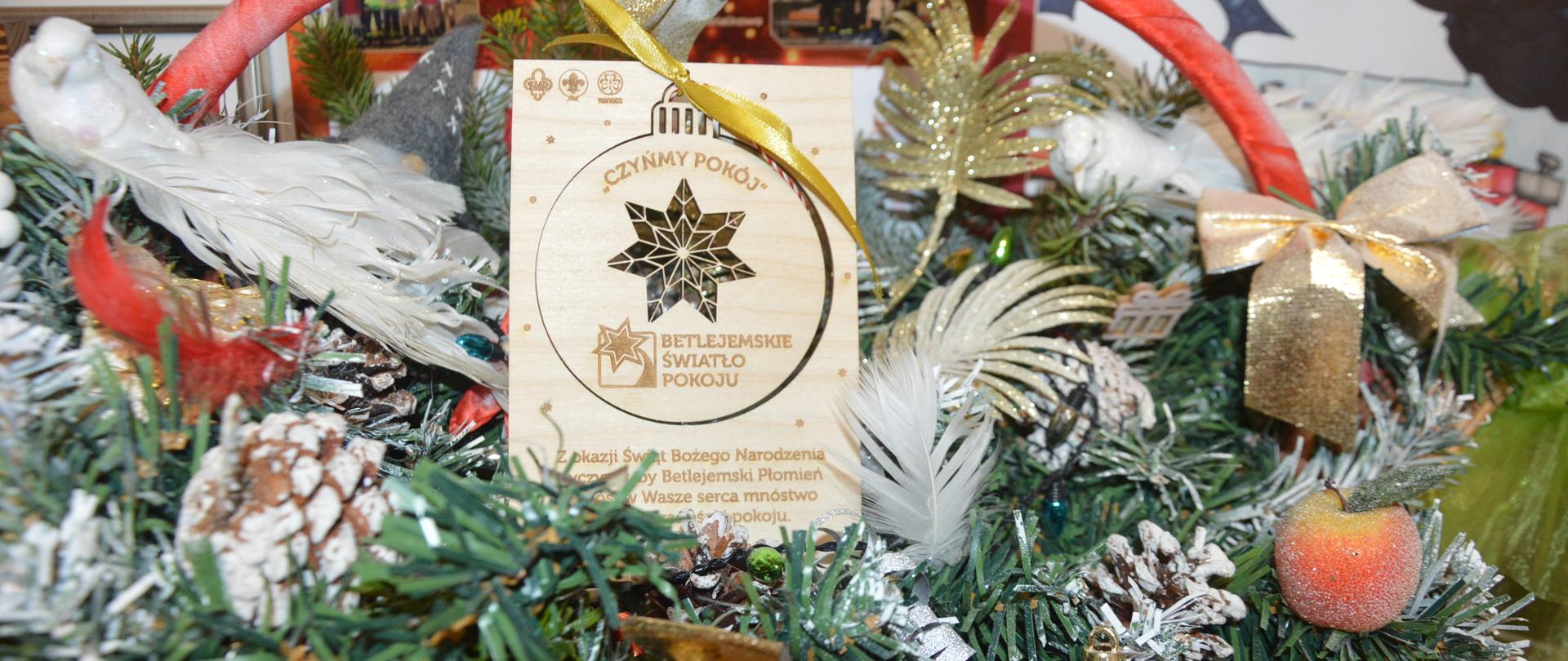 Stroik świąteczny, wykonany z gałązek świerki, bombek, wstążek i innych ozdób. W centralnym miejscu znajduje sie tabliczka z życzeniami od harcerzy.