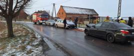 Na jednym pasie ruchu znajdują dwa samochody osobowe uczestniczące w kolizji oraz nieoznakowany radiowóz policyjny i wóz straży pożarnej. Na chodniku, wokół pojazdów przebywają strażacy i policjanci.W tle zabudowania. Z lewej strony rośnie drzewo