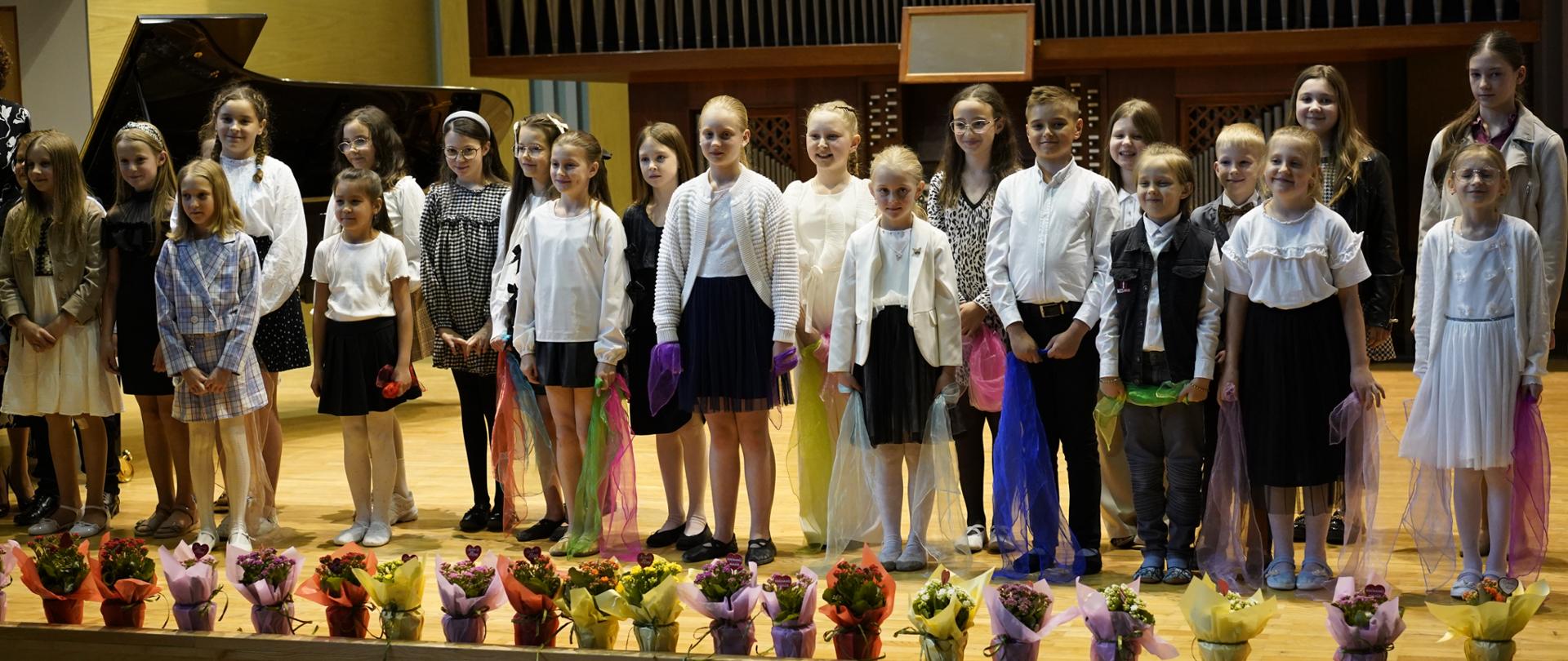 Na szerokim ujęciu widać grupę dzieci i młodzieży stojącą na scenie, w tle której znajduje się ściana z dużym, ciemnym prospektem organowym. Po lewej stronie sceny widoczny jest również czarny fortepian. Dzieci, w większości dziewczynki, są ubrane w eleganckie stroje, głównie w odcieniach bieli, czerni i granatu, niektóre z kolorowymi akcentami, takimi jak wstążki czy ozdobne szale. Stoją w luźnych rzędach, twarzami zwrócone do widza. Przed nimi, na skraju sceny, poustawiane są kolorowe bukiety kwiatów w doniczkach, prawdopodobnie przygotowane do wręczenia. Scena jest drewniana, a w tle widać niewielki kawałek widowni. Oświetlenie jest jasne i równomierne, co podkreśla szczegóły sceny i postaci.