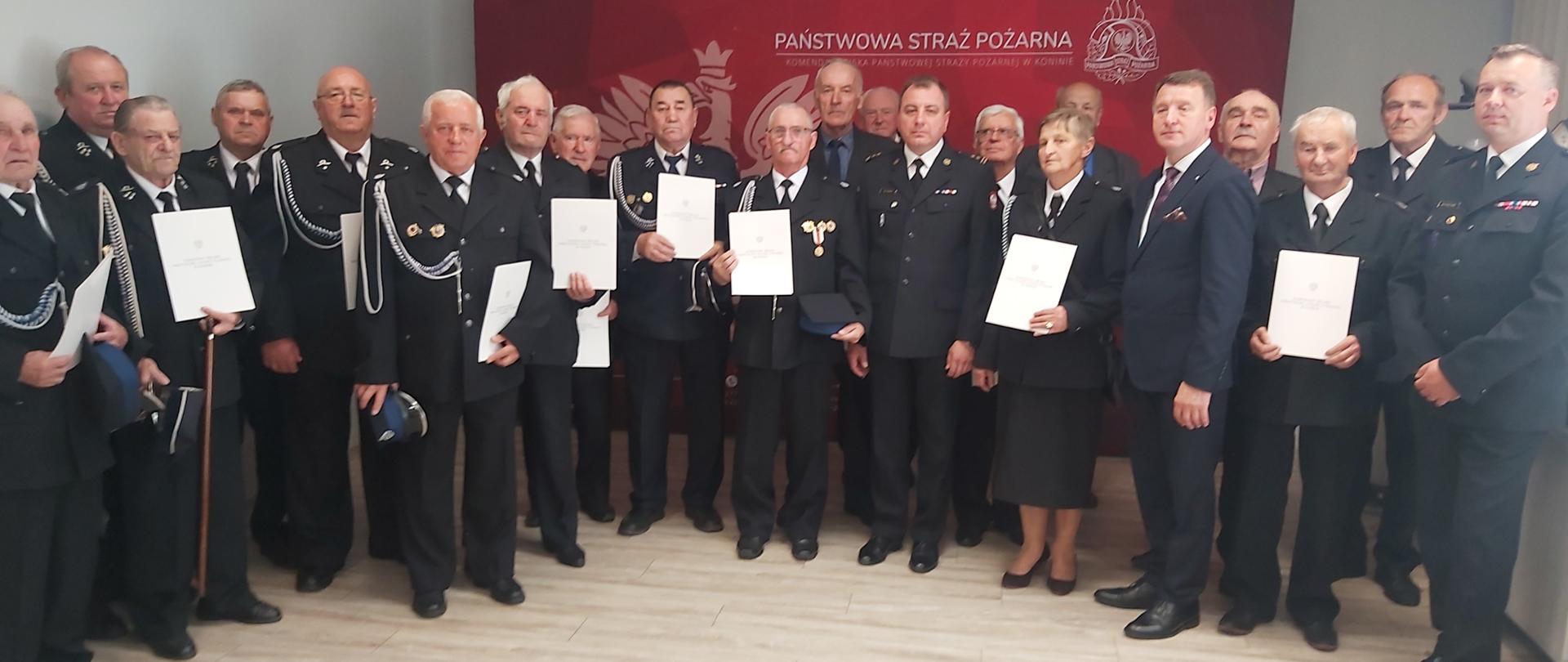 Zdjęcie przedstawia Posła na Sejm RP, komendantów oraz druhów OSP, którzy otrzymali decyzje o przyznaniu świadczenia ratowniczego. Wszyscy stoją w świetlicy przed banerem komendy.