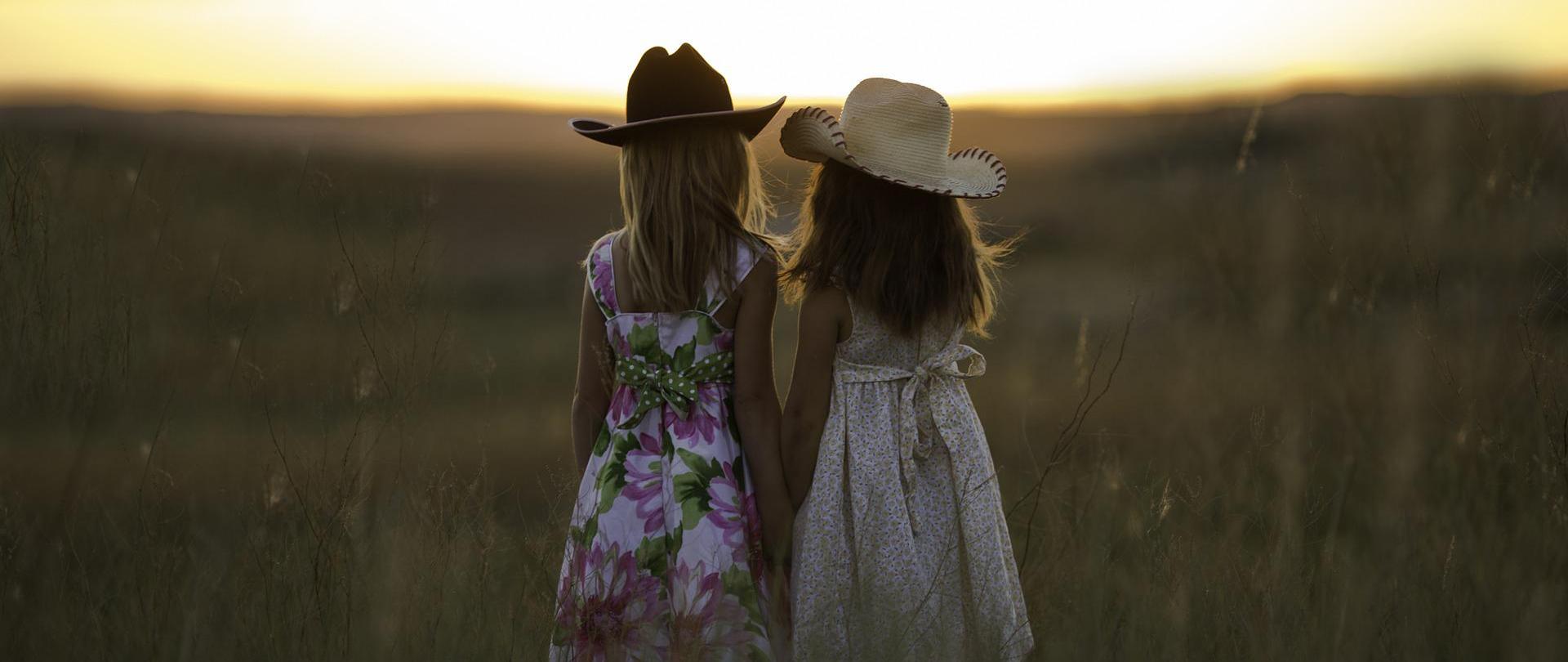 Dwie dziewczynki stojące tyłem, w letnich sukienkach, na łące, patrzące w stronę słońca