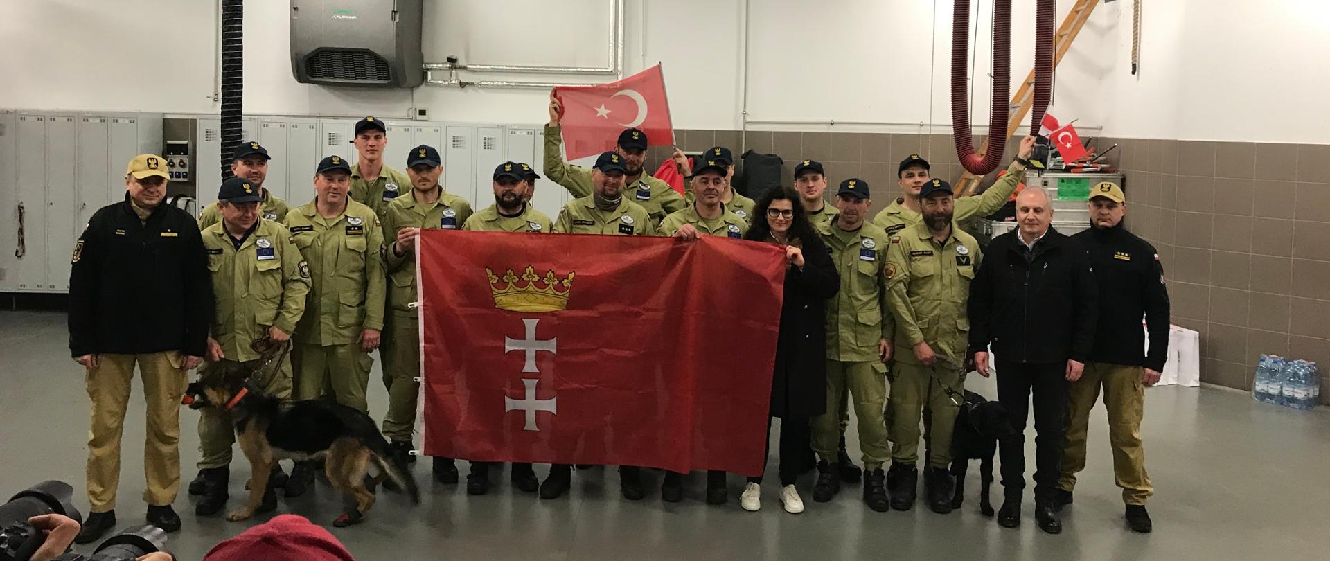 Strażacy z grupy poszukiwawczej trzymają flagę miasta Gdańska stoją wspólnie z wojewodą pomorskim pomorskim komendantem wojewódzkim Państwowej Straży Pożarnej prezydent Gdańska oraz komendantem miejskim Państwowej Straży Pożarnej w Gdańsku.