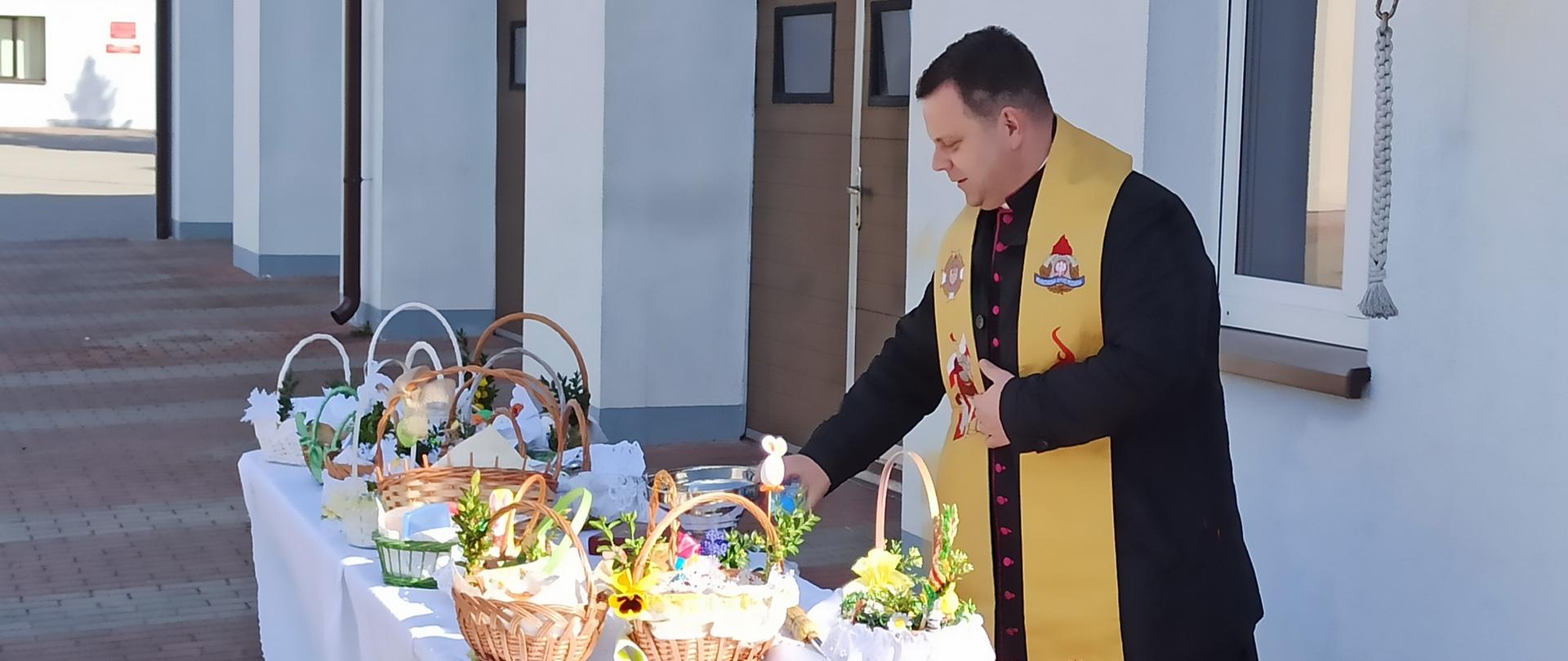 Zdjęcie przedstawia księdza kanonika Mariusza Stasiaka dziekana Dekanatu Brodnickiego, a zarazem kapelana strażaków w województwie kujawsko-pomorskim – oglądającego koszyczki z pokarmami. W tle bramy wyjazdowe Jednostki Ratowniczo-Gaśniczej.