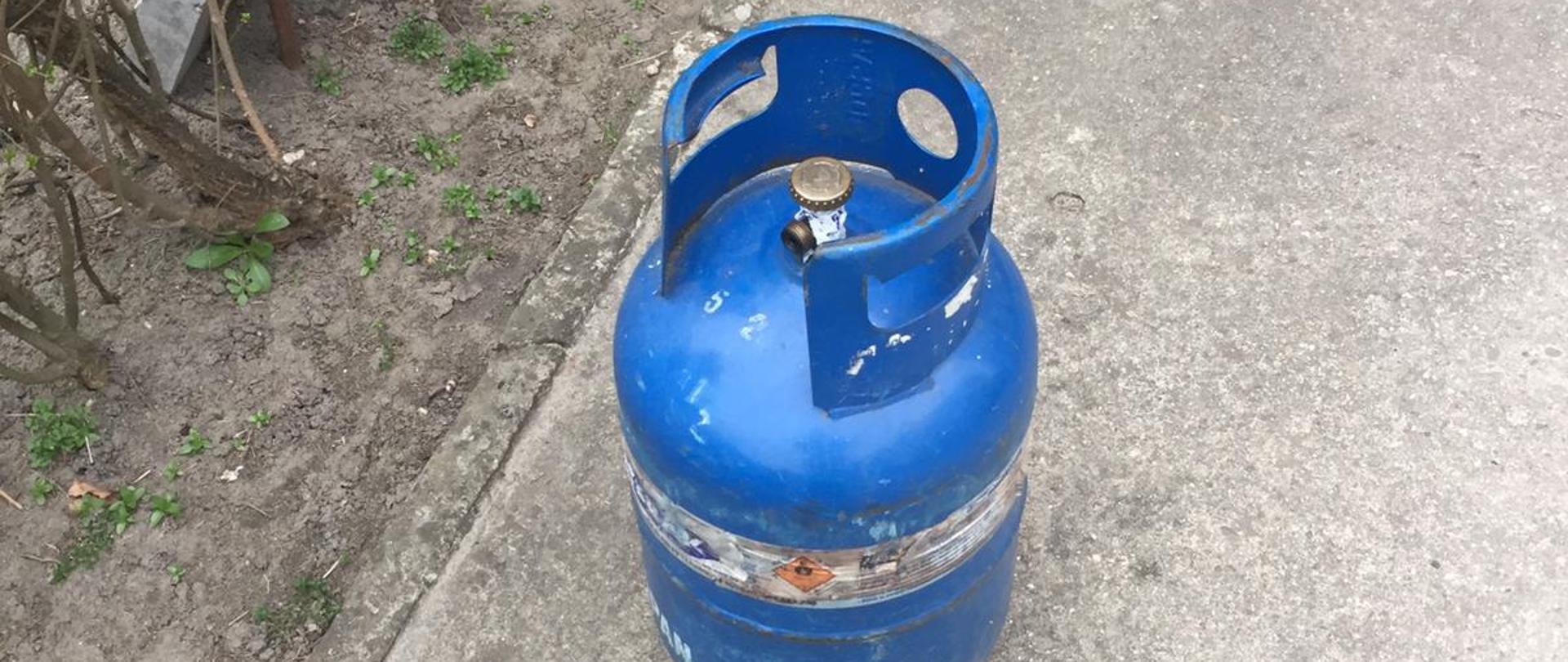Na zdjęciu widoczna niebieska butla z gazem propan butan. Stojąca przed klatką schodową. 