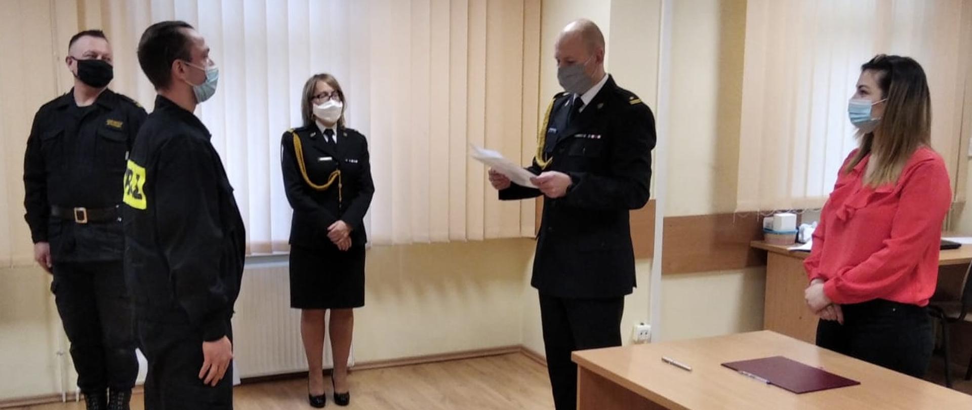 Strażak ubrany w ubranie koszarowe stoi na baczność przed komendantem, który odczytuje akt mianowania. Obok komendanta stoi kobieta - pracownik cywilny w czerwonej bluzce, czarnych spodniach i w masce, a funkcjonariuszka ubrana w mundur wyjściowy obok której stoi dowódca uroczystości ubrany w ubranie dowódczo sztabowe. 