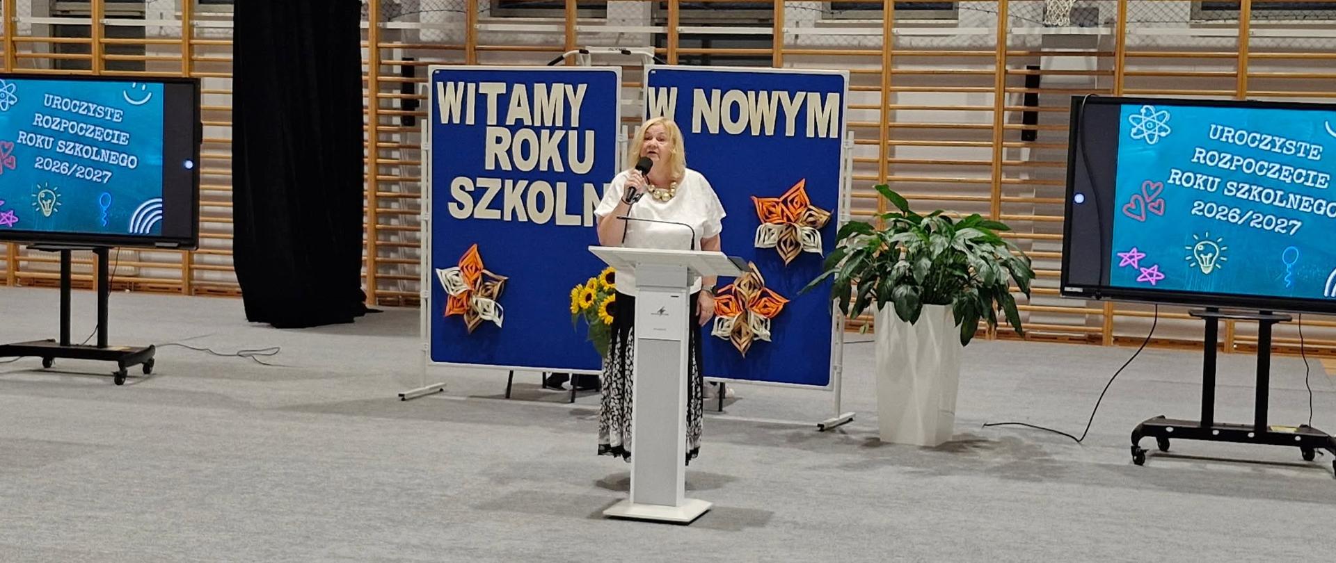 kobieta przemawia do mikrofonu, w tle, w środkowej części zdjęcia baner z napisem Witamy Nowy Rok Szkolny , po prawej i lewej stronie ekrany telewizyjne z napisem: Uroczyste rozpoczęcie roku szkolnego 2026/2027