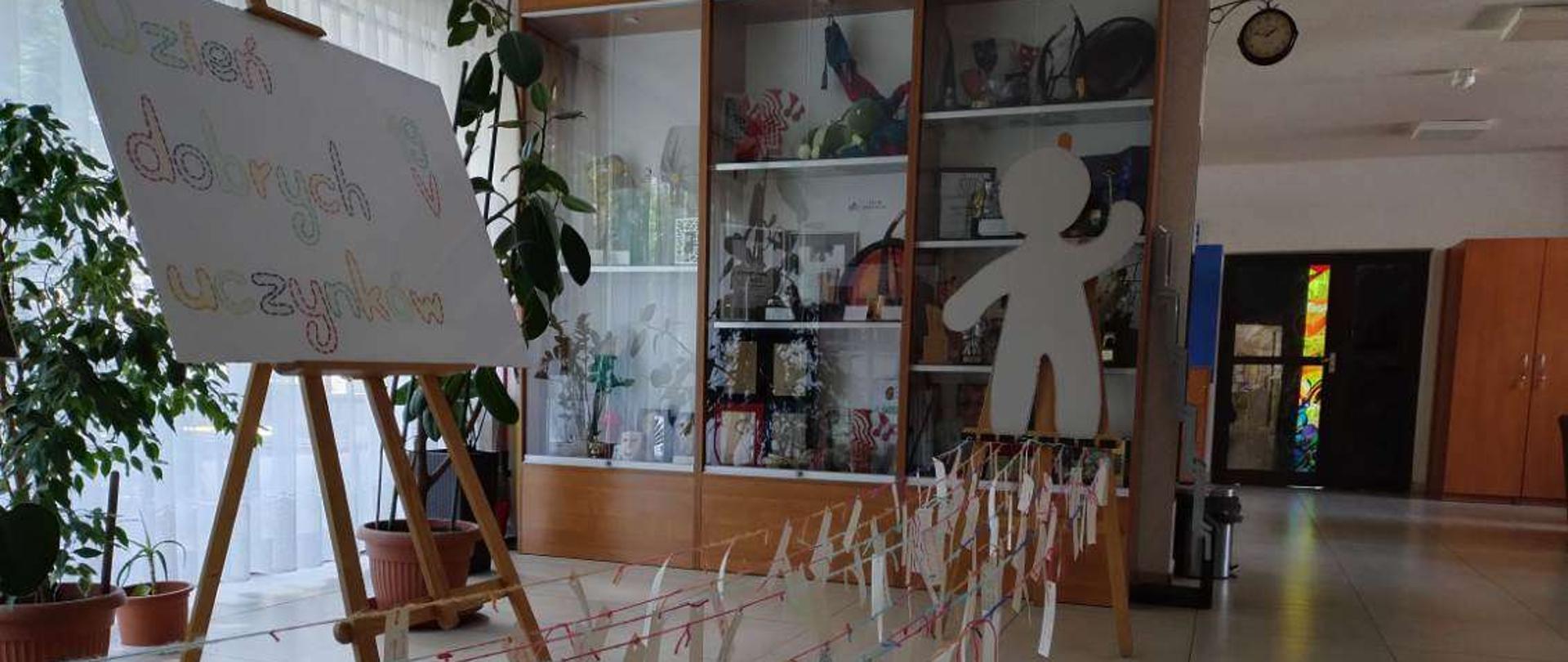 W holu po lewej stronie na tle okna stoi kilka dużych roślin w donicach, a przed nimi drewniana sztaluga z dużym białym plakatem, a nim kolorowy napis: "Dzień dobrych uczynków 19 V". Na środku w tle widać oszkloną gablotę. Przed gablota na sztaludze jest umieszczony duży tekturowy ludzik, a przed nim w lewą stronę rozciągniętych jest kilka kolorowych włóczek, do których są zaczepione karteczki z kolorowymi napisami. Po prawej stronie w tle widać fragment szafy i drzwi.