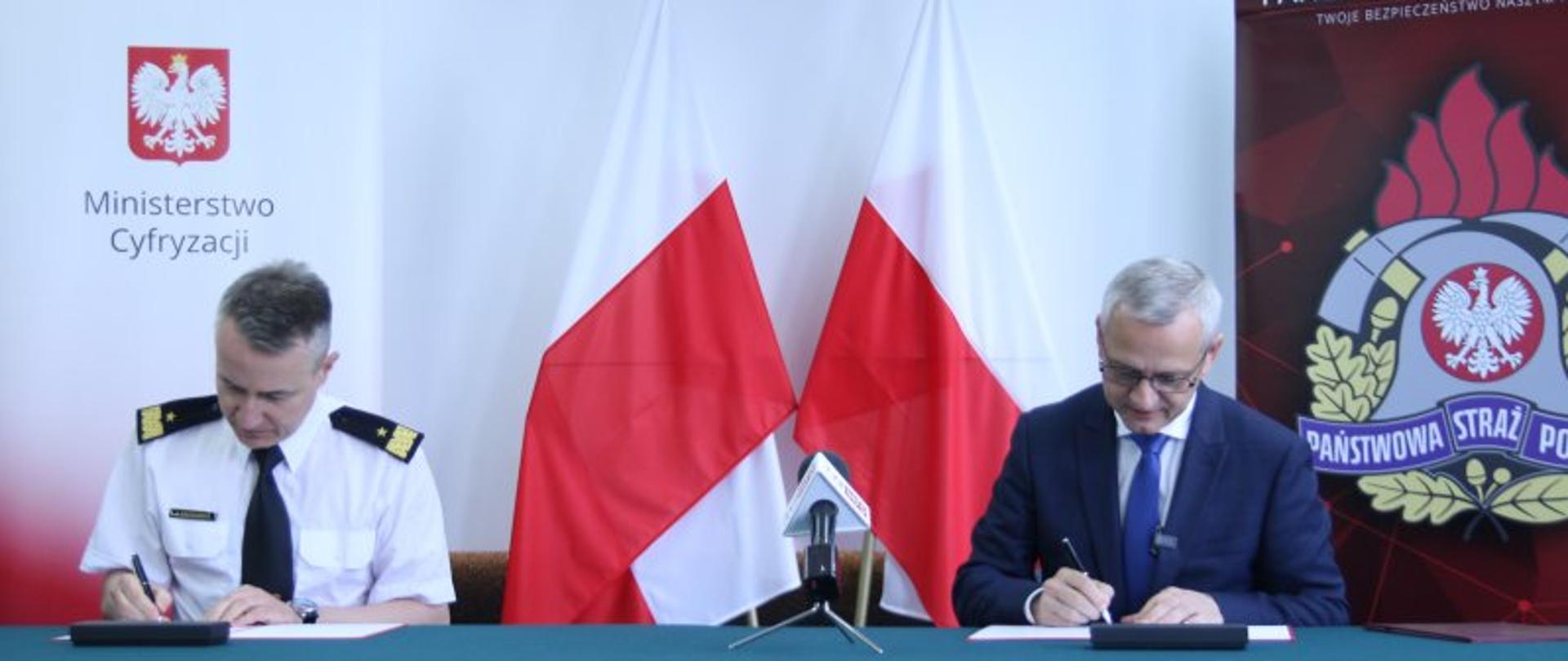 Podpisanie porozumienia pomiędzy Komendantem Głównym Państwowej Straży Pożarnej a Ministerstwem Cyfryzacji – na zdjęciu moment podpisywania Porozumienia na tle biało-czerwonych flag. Z boku widoczne logotypy MC i PSP