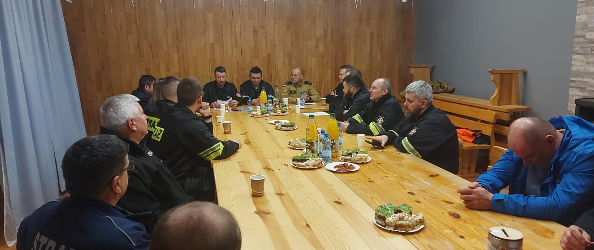 Na zdjęciu widać strażaków OSP KSRG z Wojtkowej oraz komendanta powiatowego PSP z Ustrzyk Dolnych. Wszyscy siedzą przy stolena którym są napoje i przekąski. Zdjęcie wykonane w środku pomieszczenia.