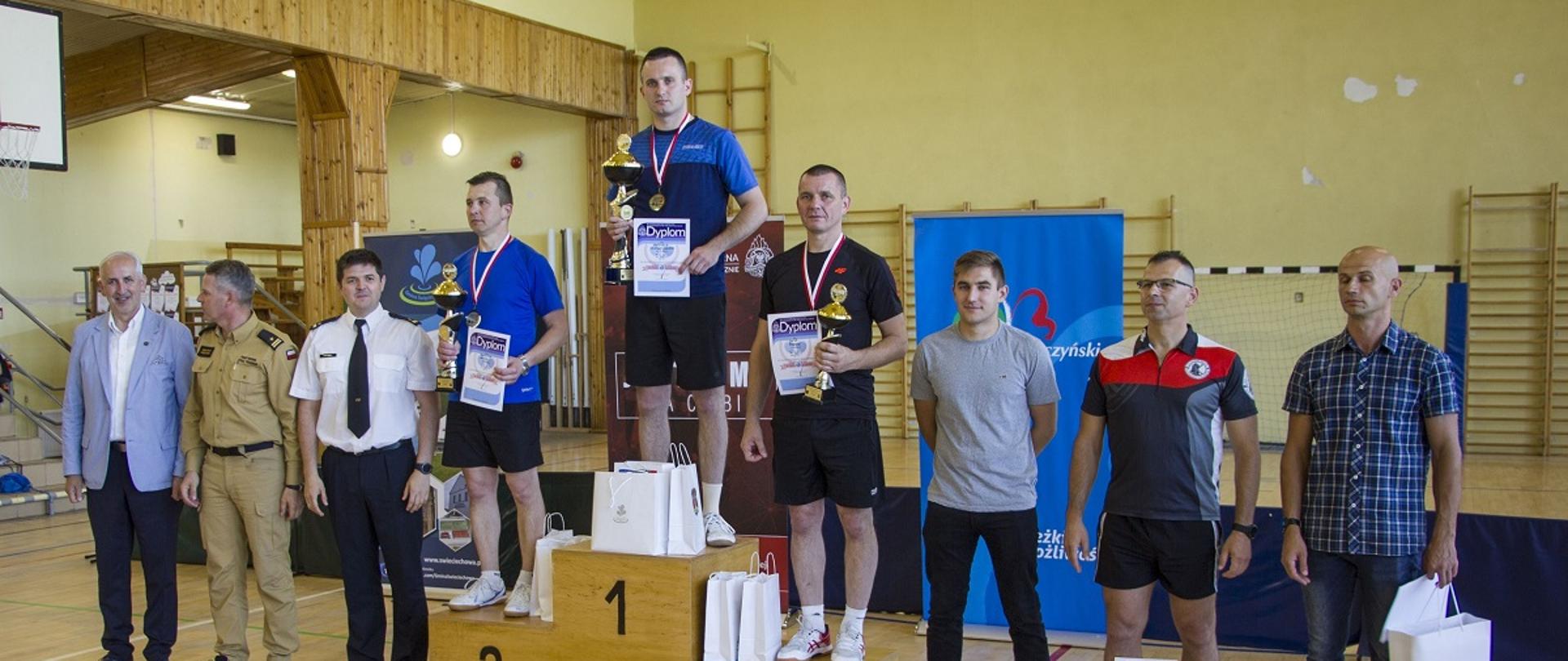 grupa mężczyzn stoi obok siebie, pomiędzy nimi podium, na ktorym stoją sportowcy