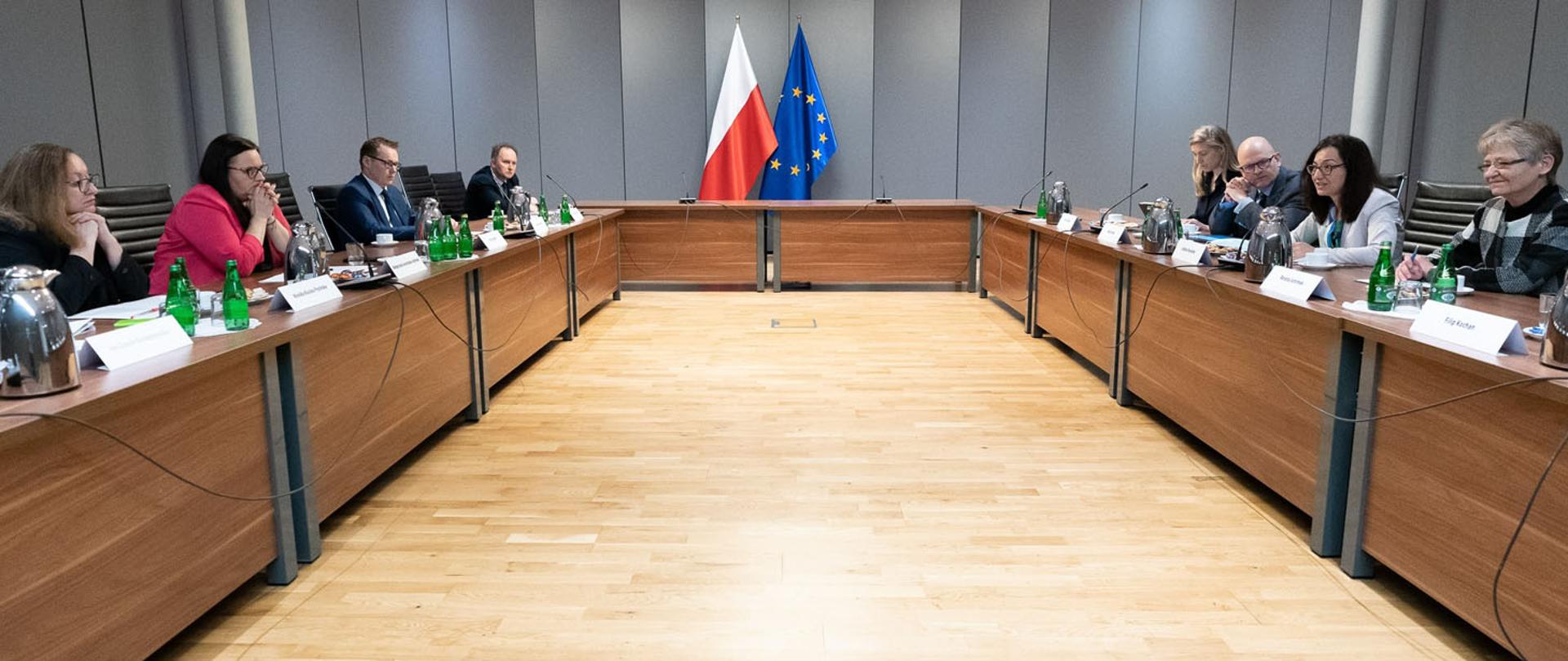 W sali konferencyjnej grupa osób siedzi przy stołach ustawionych w podkowę. Przy ścianie dwie flagi PL i UE.