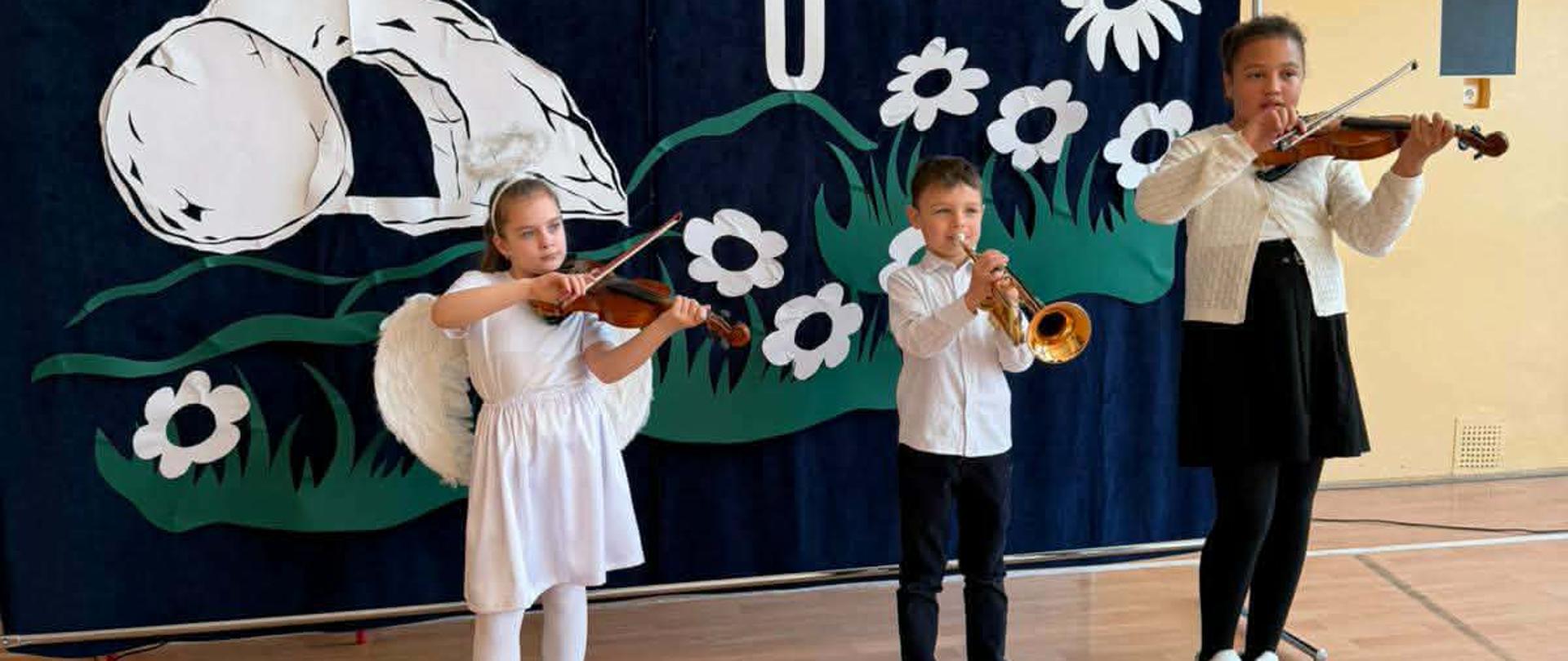 Na zdjęciu widoczna jest trójka dzieci występujących na scenie podczas uroczystości wielkanocnej. Wydarzenie odbywa się na tle ciemnoniebieskiej kurtyny z dekoracjami i napisem w języku polskim. Tło stanowi duża płachta z napisem: „WIELKANOC 2026 OD CISZY DO ALLELUJA”. Na dekoracji znajdują się białe elementy symboliczne: Pusty grób z odsuniętym kamieniem po lewej stronie. Krzyż umieszczony centralnie. Białe kwiaty i zielona trawa u dołu dekoracji. Wykonawcy i instrumenty Na pierwszym planie stoją dzieci grające na instrumentach: Po lewej stronie: Dziewczynka ubrana w białą sukienkę, białe rajstopy i buty. Ma na sobie skrzydła anioła oraz aureolę. Gra na skrzypcach. W środku: Chłopiec w białej koszuli i ciemnych spodniach, który gra na trąbce. Po prawej stronie: Starsza dziewczynka ubrana w biały sweterek, czarną spódnicę i czarne leginsy. Ona również gra na skrzypcach. Dzieci stoją na jasnej, drewnianej podłodze (prawdopodobnie parkiet w sali gimnastycznej lub świetlicy), a cała scena jest oświetlona jasnym, naturalnym światłem. Atmosfera zdjęcia sugeruje szkolne lub przedszkolne przedstawienie z okazji świąt.