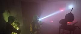 Zdjęcie przedstawia dwóch strażaków w zadymionym pomieszczeniu w aparatach ochrony układu oddechowego i z włączonymi latarkami. Obok nich w pomieszczeniu stoi maszyna.