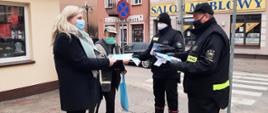 Zdjęcie przedstawia dwóch strażaków OSP, którzy wręczają kobiecie i mężczyźnie ulotki dotyczące szczepień przeciwko COVID-19. Zdjęcie wykonane jest w mieście, a osoby te znajdują się na chodniku obok sklepu. W tle widać inne sklepy, samochód oraz znaki drogowe. 
