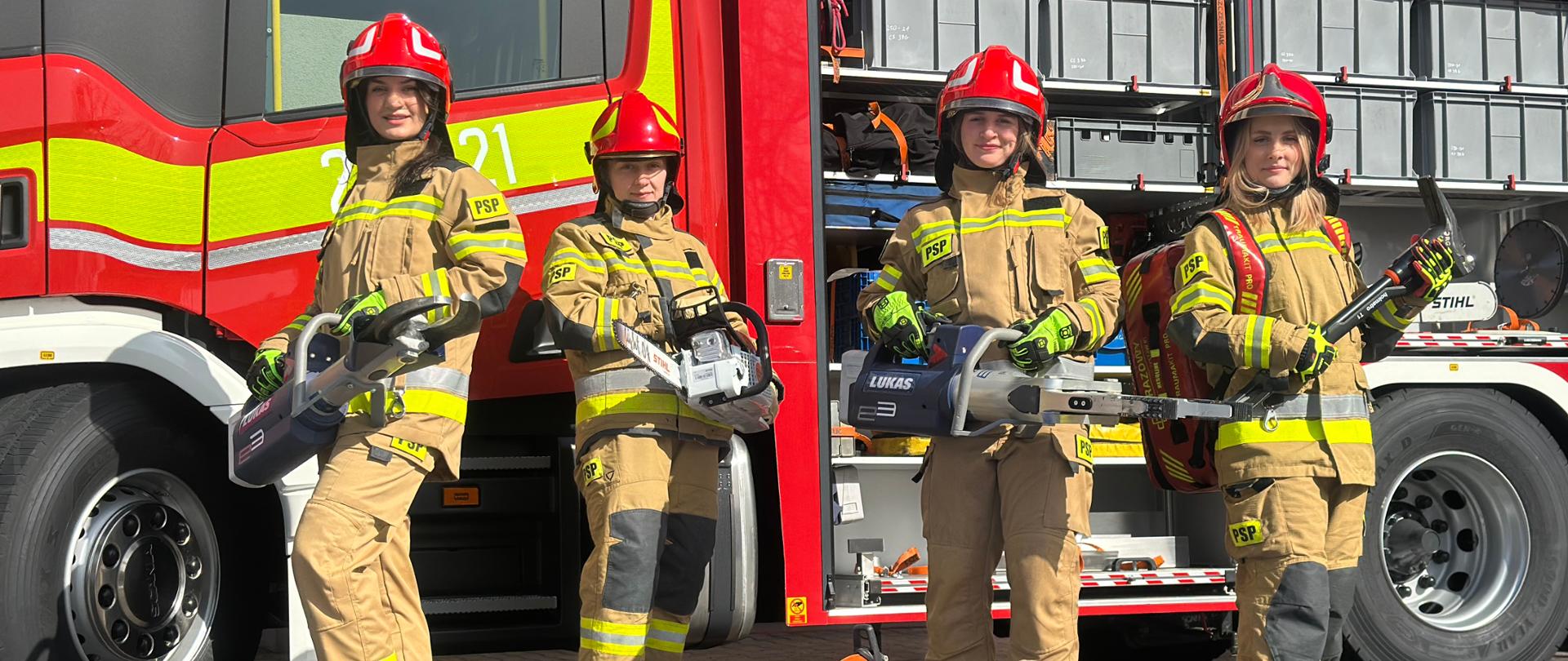 Cztery strażaczki w ubraniach specjalnych stoją przed otwartym samochodem ratowniczo-gaśniczym. Każda z nich trzyma sprzęt wykorzystywany podczas działań ratowniczych, m.in. pilarki i narzędzia hydrauliczne. Na pierwszym planie na macie leżą dodatkowe elementy sprzętu ratowniczego. W tle widoczne są skrytki sprzętowe wozu strażackiego