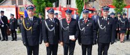 Zdjęcie przedstawia czterech strażaków w mundurach Ochotniczej Straży Pożarnej oraz Komendanta Powiatowego PSP w Ciechanowie stojących w szeregu na tle wozów strażackich i uczestników uroczystości. Środkowy strażak trzyma odznaczenie i ma na szyi medal, co sugeruje, że właśnie został uhonorowany. W tle widoczni są inni strażacy, kobiety i mężczyźni, również w mundurach, a także flagi i banery jednostek OSP. Całość ma charakter oficjalnej ceremonii.