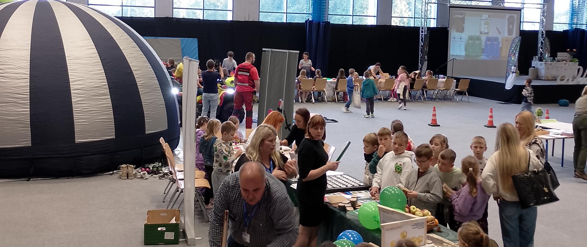 Zdjęcie przedstawia halę sportową wypełnioną stanowiskami różnych organizacji. Przy stanowisku WIORiN stoi liczna grupa dzieci. Pracownicy WIORiN prowadzą warsztaty i gry edukacyjne związane z ekologią i rolnictwem.