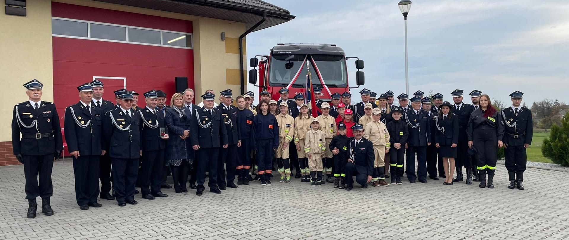 Na tle samochodu pożarniczego oraz remizy strażackiej stoi grupa ludzi, w większości strażacy w umundurowaniu galowym oraz Młodzieżowa Drużyna Pożarnicza.