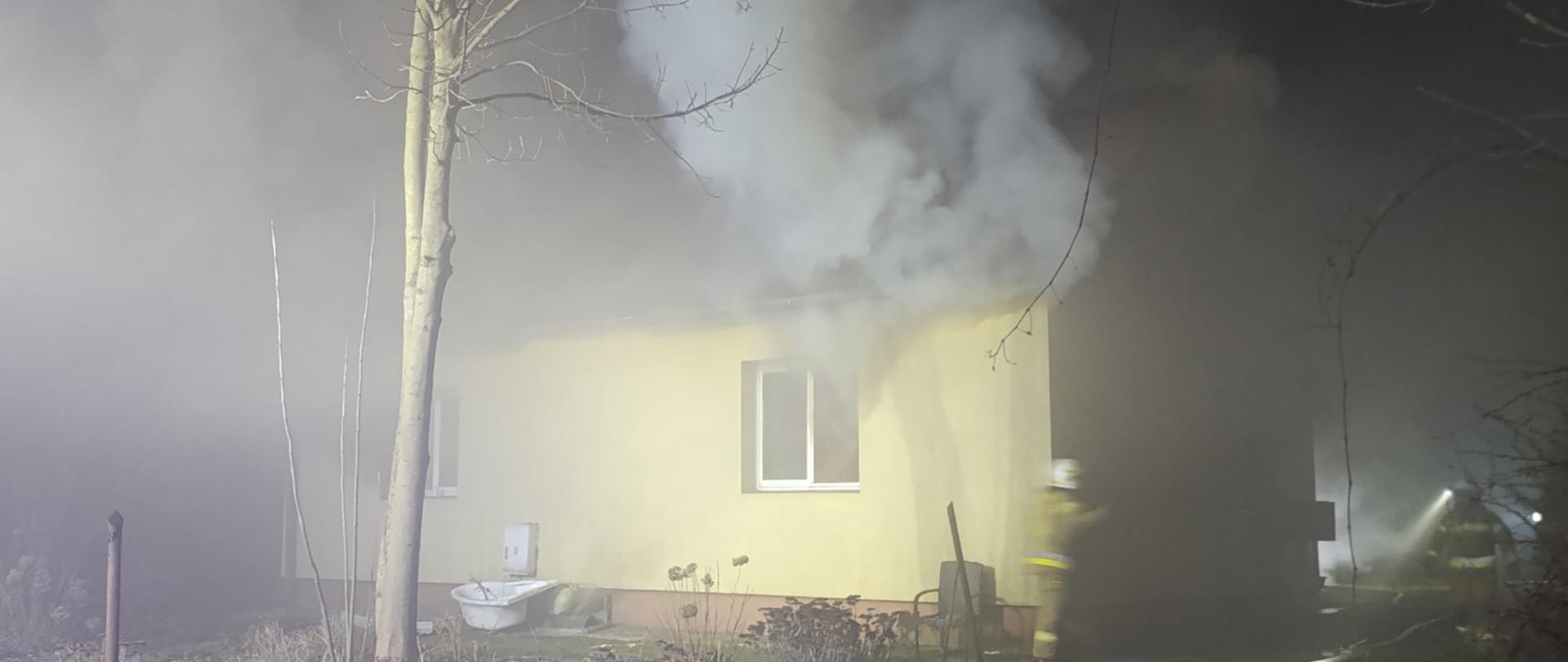 Zdjęcie przedstawia parterowy, murowany budynek mieszkalny, z którego przez jedno z okien wydobywa się dym. Obok domu widoczna jest rozwinięta linia gaśnicza, a także dwóch strażaków.