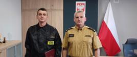 Komendant Powiatowy Państwowej Straży Pożarnej w Człuchowie st. bryg. Radosław Batruch stoi w gabinecie i wręcza akt mianowania na stanowisko dowódcy zmiany st. asp. Karolowi Płóciennik.