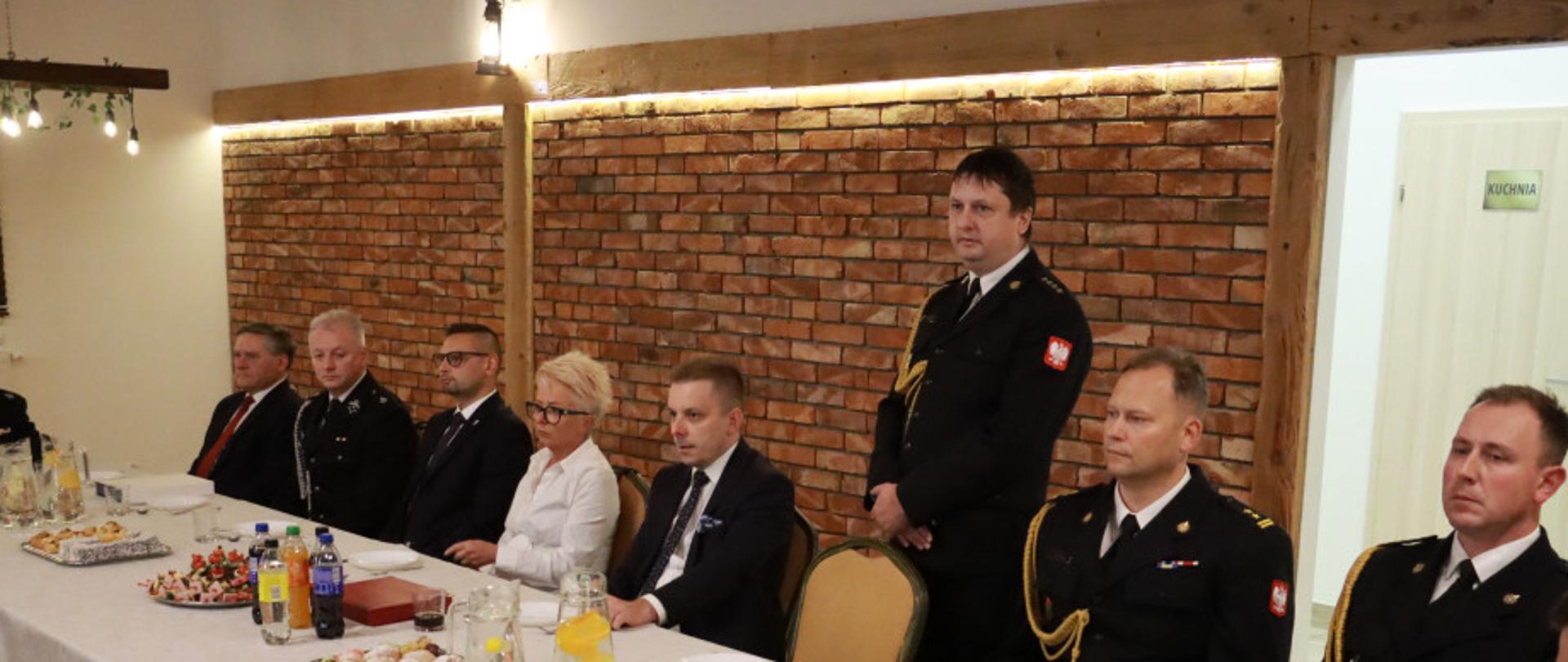 Uczestnicy spotkania siedzą przy długim stole nakrytym białym obrusem. Wśród obecnych znajdują się przedstawiciele Państwowej Straży Pożarnej oraz władz samorządowych. Na stole ustawiono półmiski z przekąskami i napoje. Spotkanie odbywa się w sali o ciepłym oświetleniu i ceglanej aranżacji wnętrza.