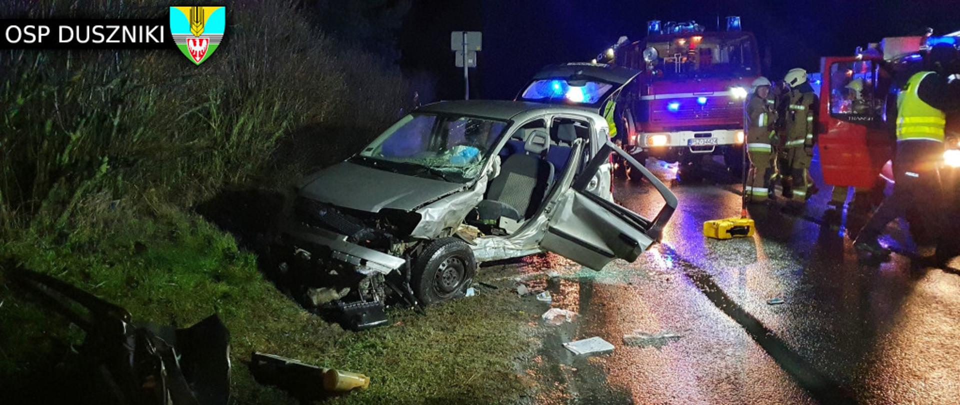 Widać uszkodzony samochód, od strony kierowcy uszkodzone drzwi, jest ciemno, po prawej jest samochód strażacki i kilku strażaków