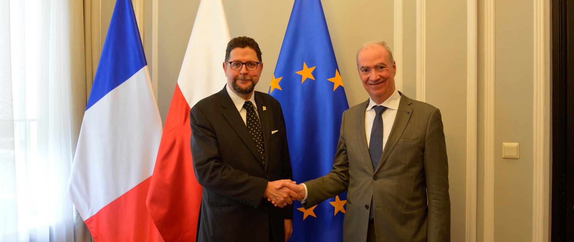 Wiceminister Szeptycki stoi obok mężczyzny w szarym garniturze, podają sobie ręce, za nimi pod ścianą flagi Polski, Francji i UE.